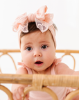 Baby wearing a pink headband with a bow, sitting behind a wooden crib.