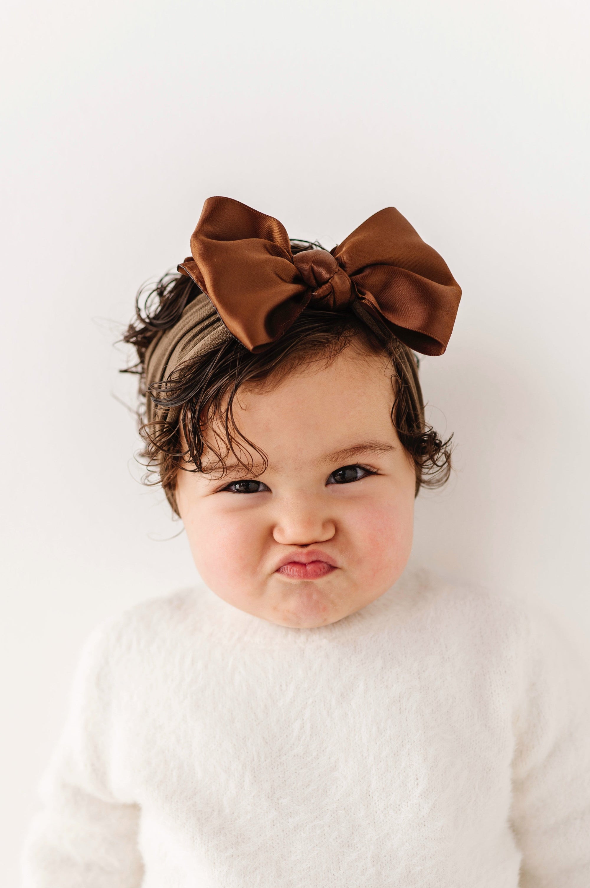 Baby wearing a white outfit with a brown bow in hair against a white background
