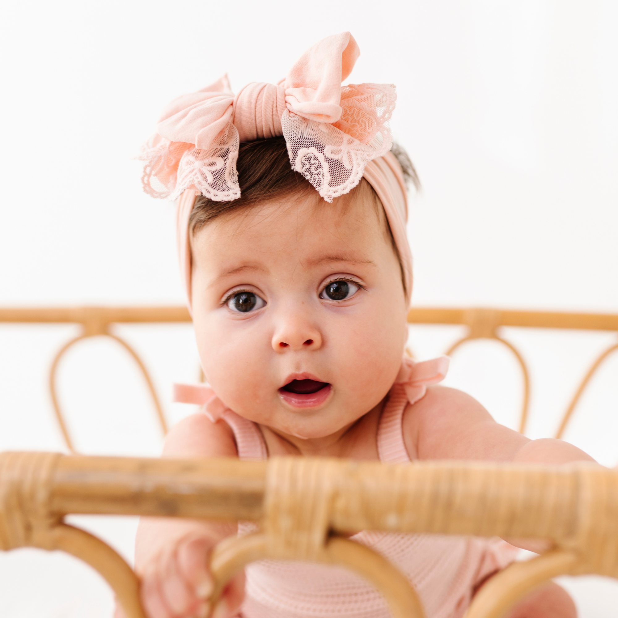 Baby wearing a pink headband with a bow, sitting behind a wooden crib.